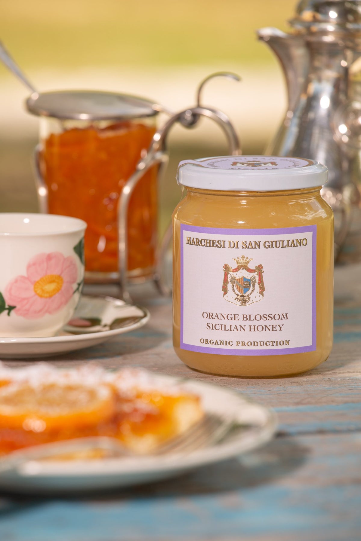 Jar of orange blossom honey on a table alongside a slice of citrus tart, a floral teacup and marmalade