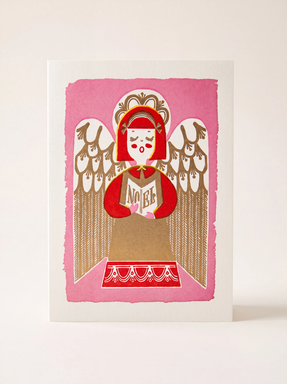 Paper greeting card with an illustrated singing angel on pink, holding a songbook that reads 'NOEL