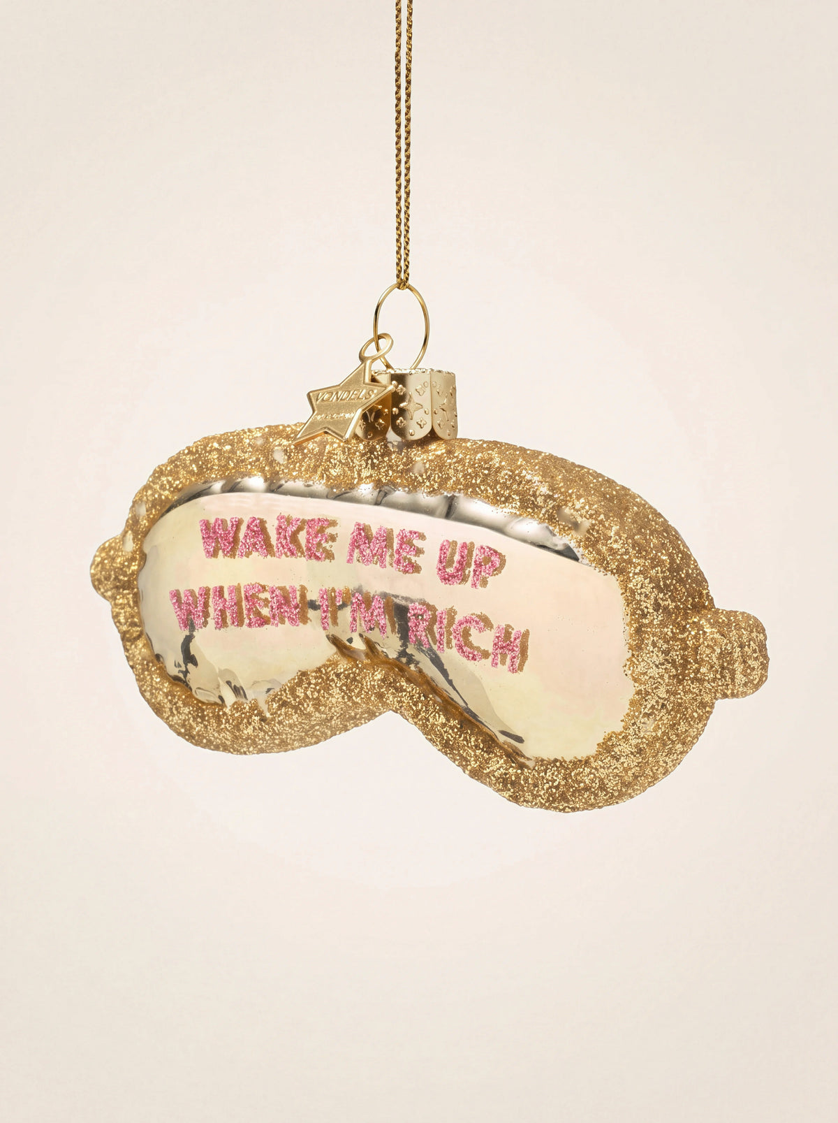 Glass sleeping mask ornament with gold glitter trim and pink lettering reading "Wake me up when I'm rich