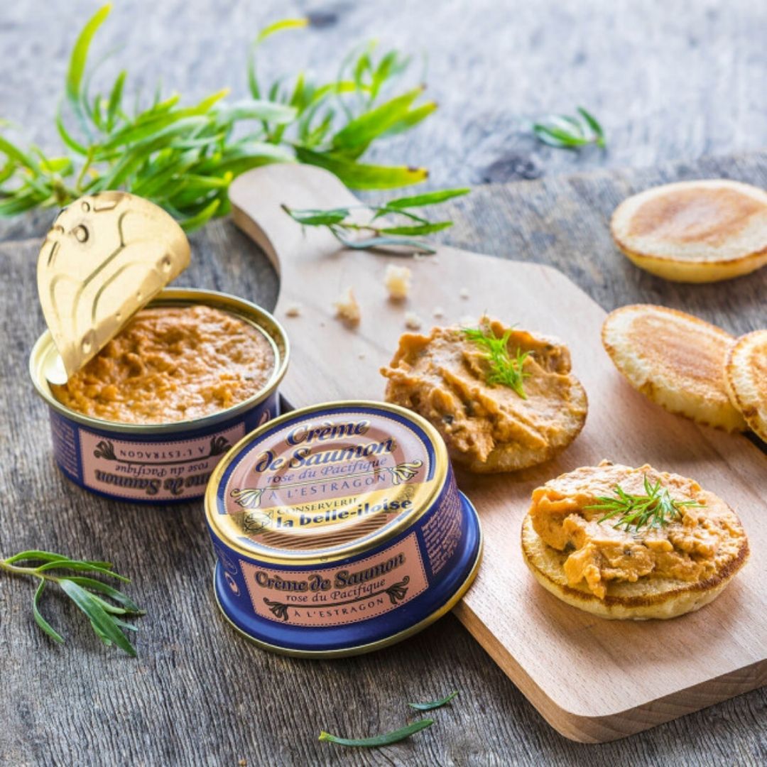 Opened tin of salmon spread alongside blinis topped with spread and fresh tarragon on a board