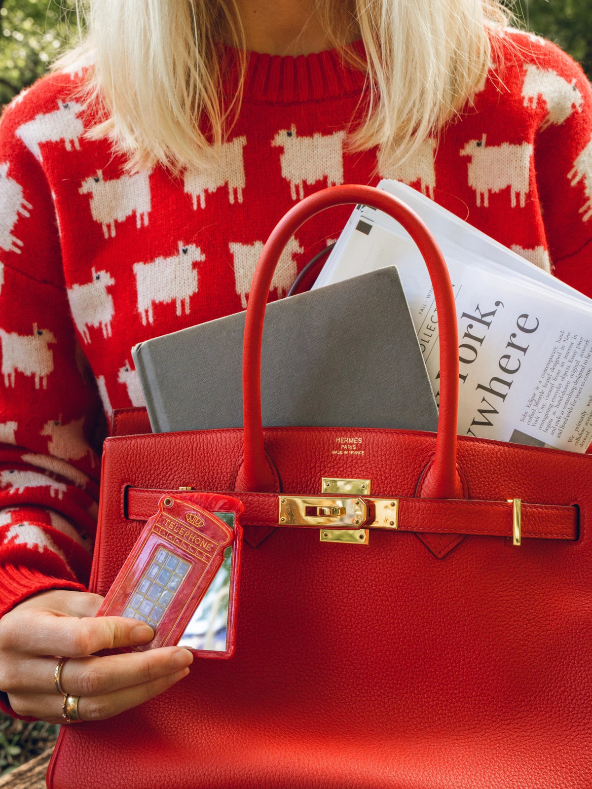 Telephone box compact mirror held swivelled open in one hand to show the mirror glass next to a red handbag