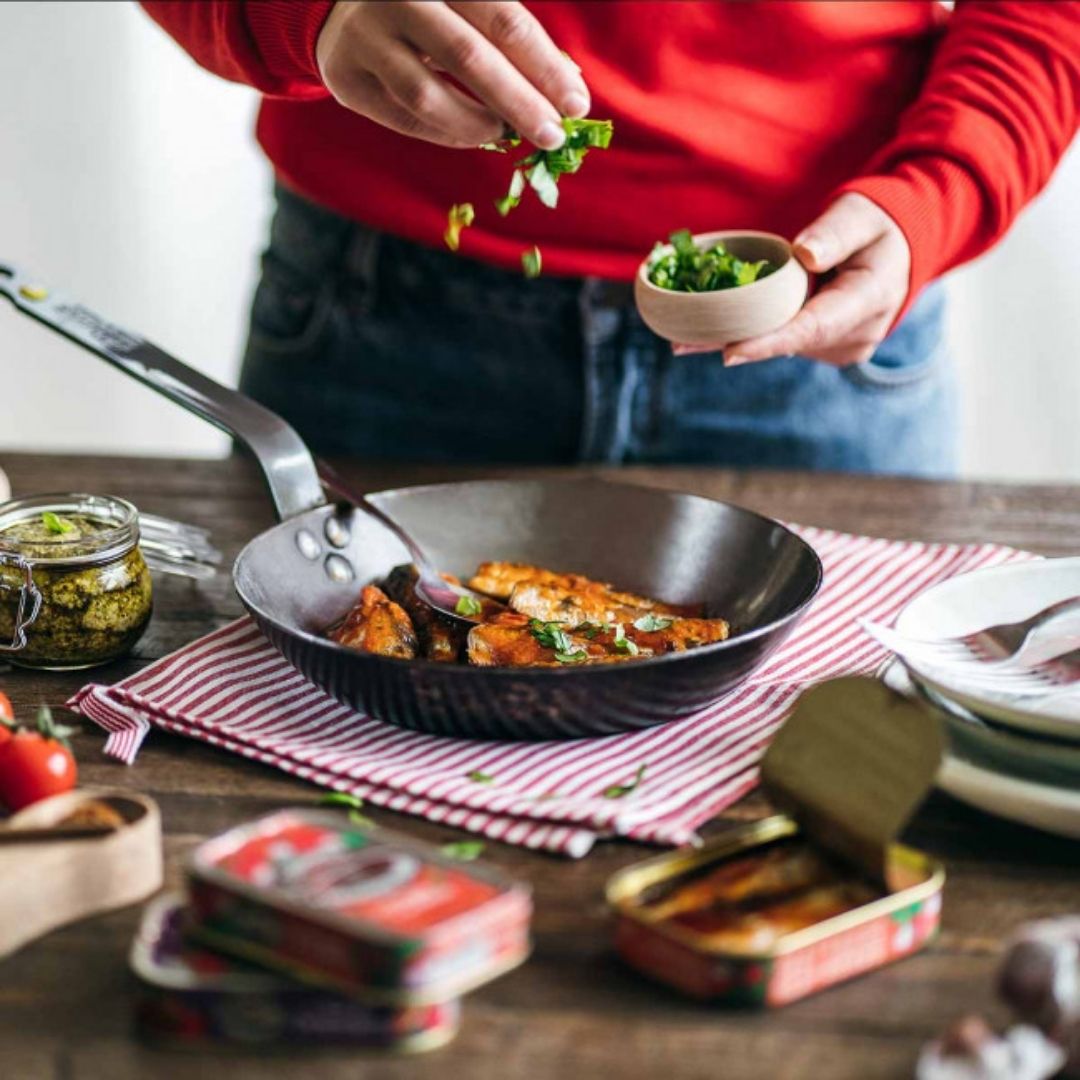 Fresh herbs sprinkled over sardines heating in a skillet beside an open tin