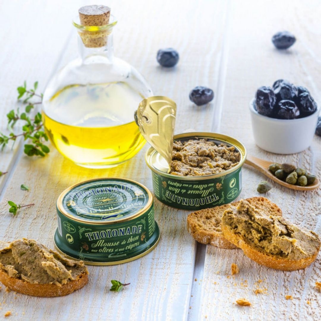Open tin showing the textured tuna mousse spread onto toasted baguette, served beside olives and capers