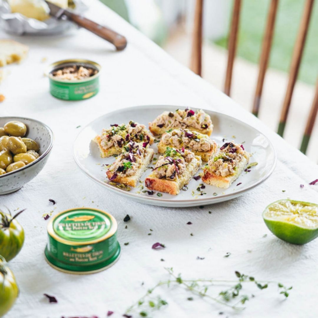 Tuna rillettes served on toasted bread slices alongside round green tins, olives and lime