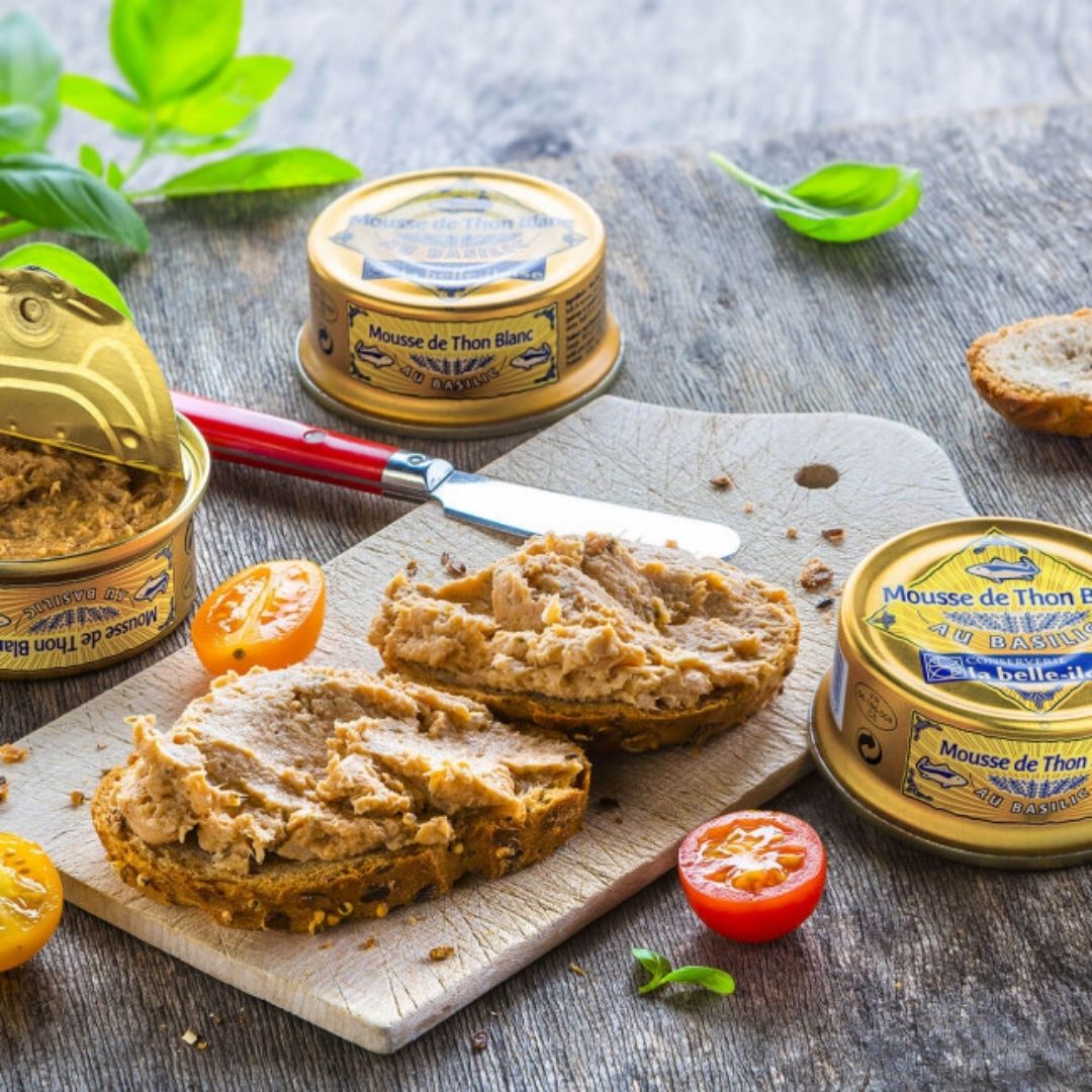 White tuna mousse spread on sliced bread next to an open tin, spreader knife and tomatoes