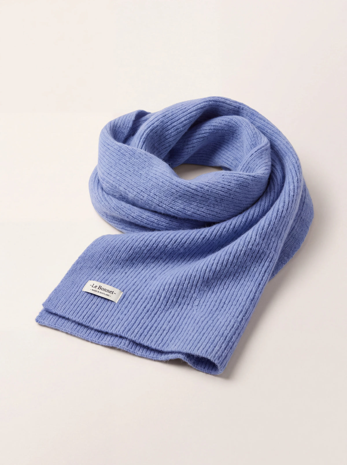 Rib-knitted light blue wool scarf loosely coiled, with a woven -Le Bonnet- label at the finished edge