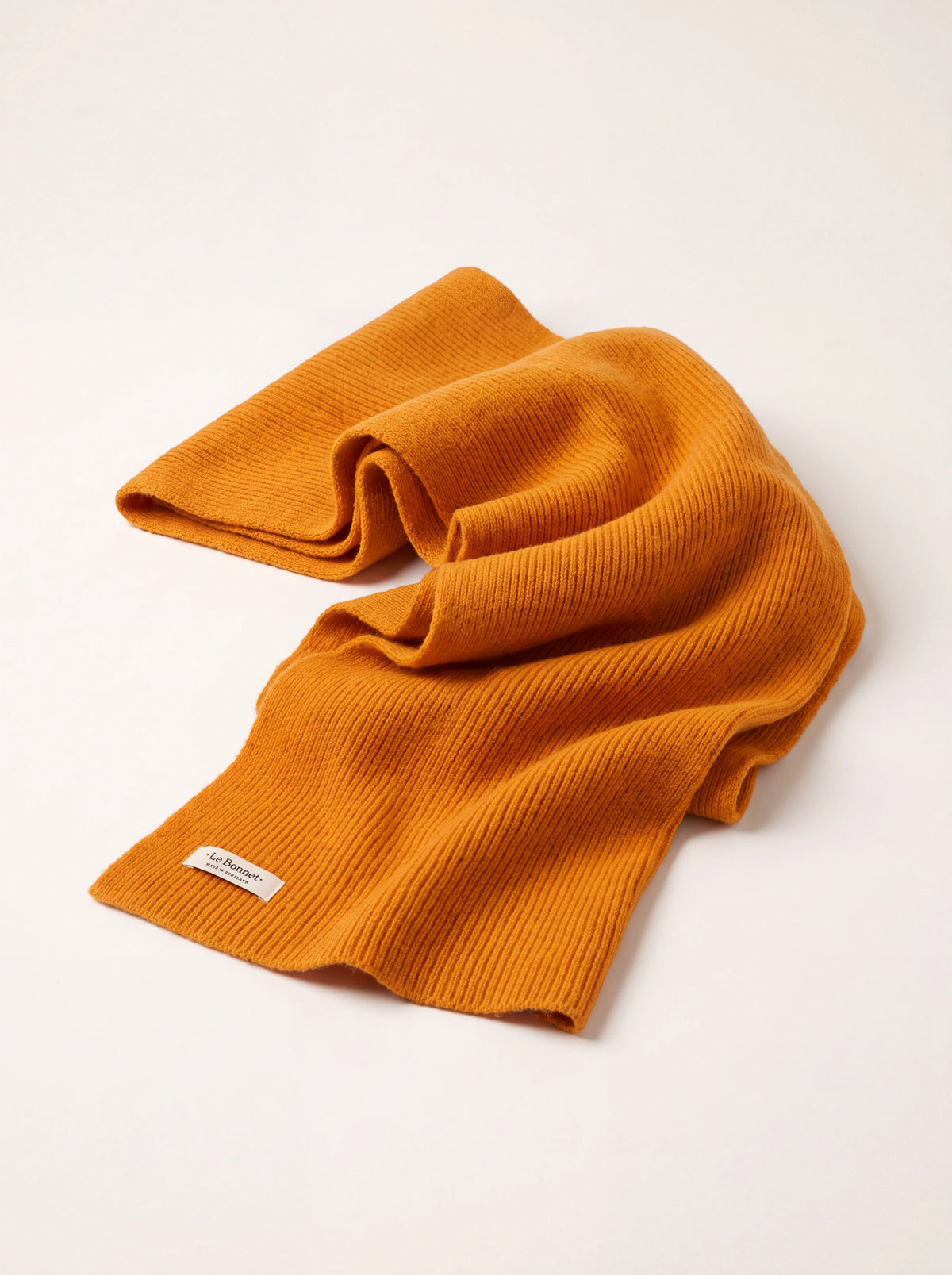 Ribbed knit wool scarf in bright orange, draped loosely with a white label reading Le Bonnet