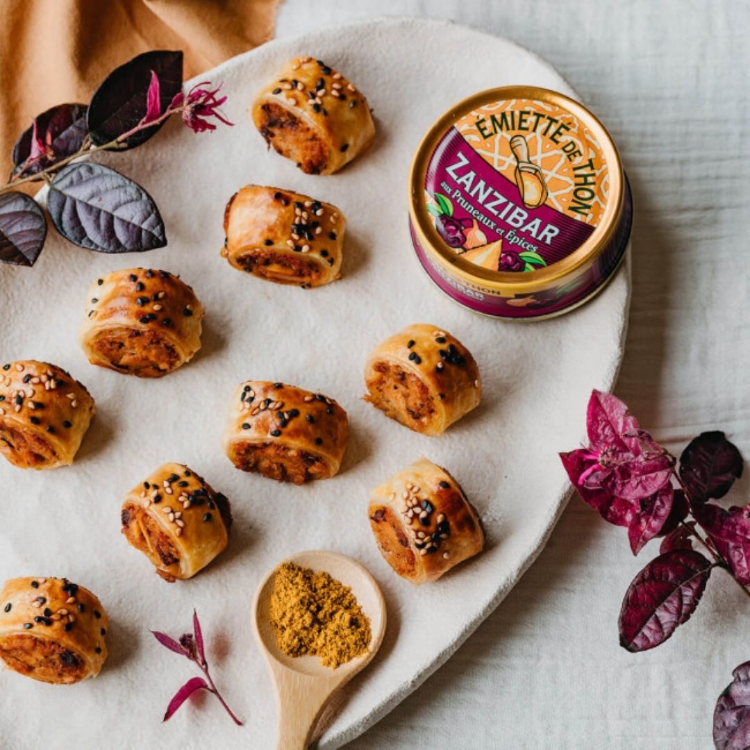 Baked savoury pastry rolls filled with flaked tuna on a platter beside the tin and a spoonful of spice
